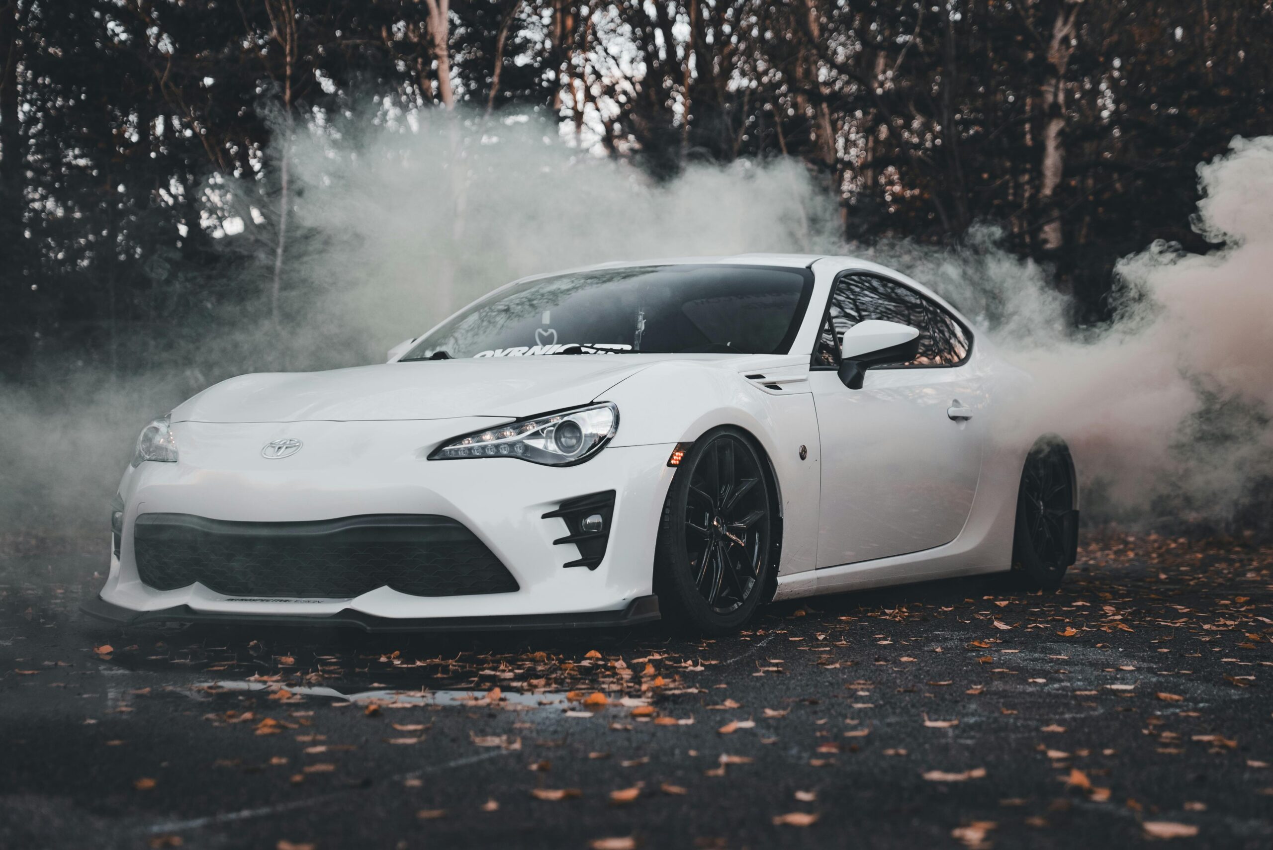 A sleek white sports car drifting through smoke on a leaf-covered road in autumn.